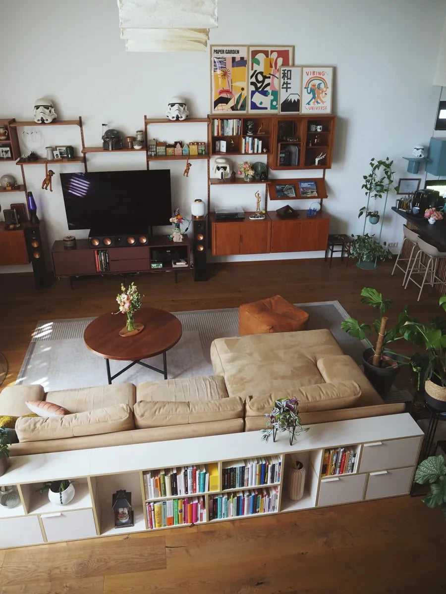 Cozy modern living room with a beige sofa, wall shelves, houseplants, and a white bookcase filled with books.